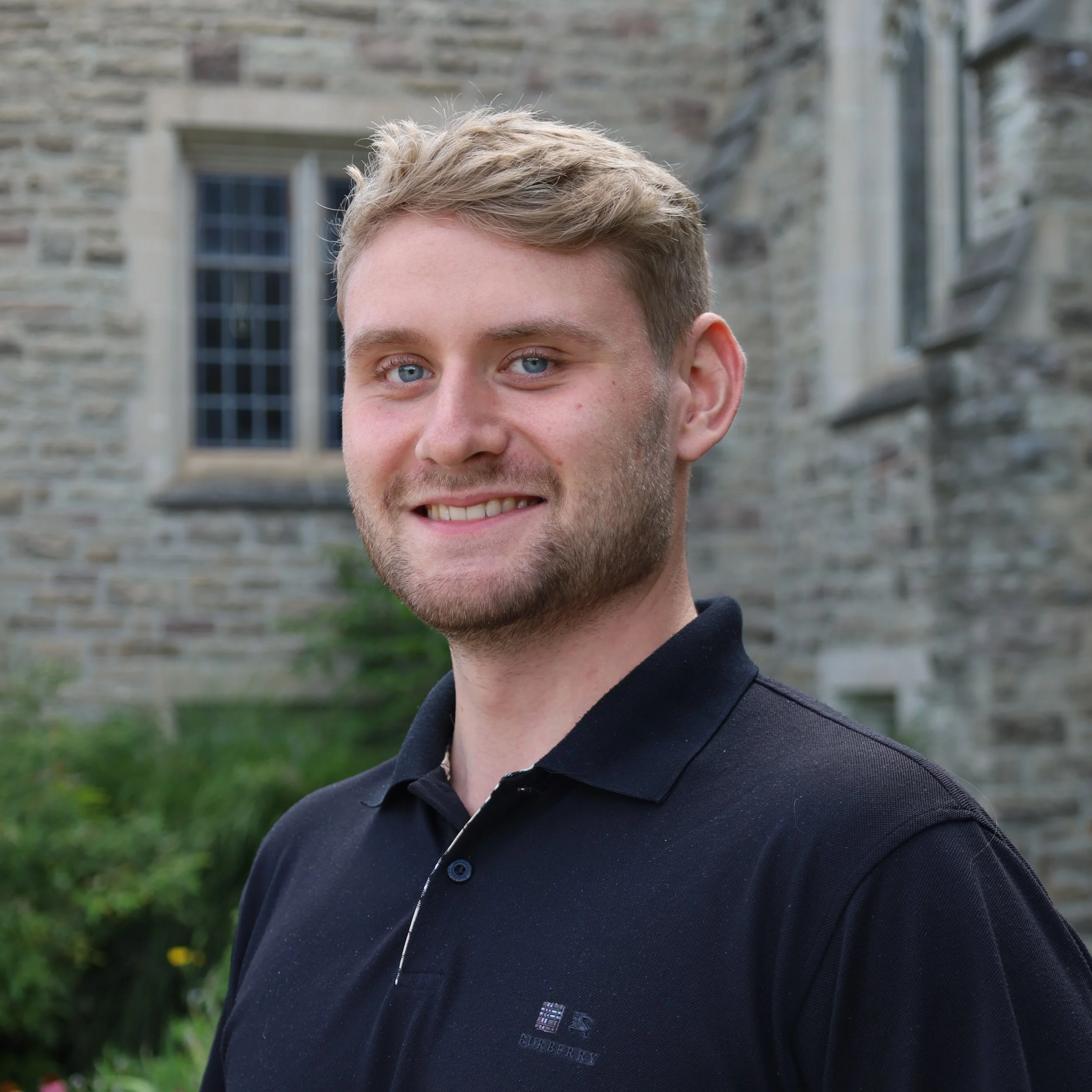 A smiling young man with blonde hair and a beard, wearing a navy polo shirt, standing outdoors in front of a stone building with greenery.
