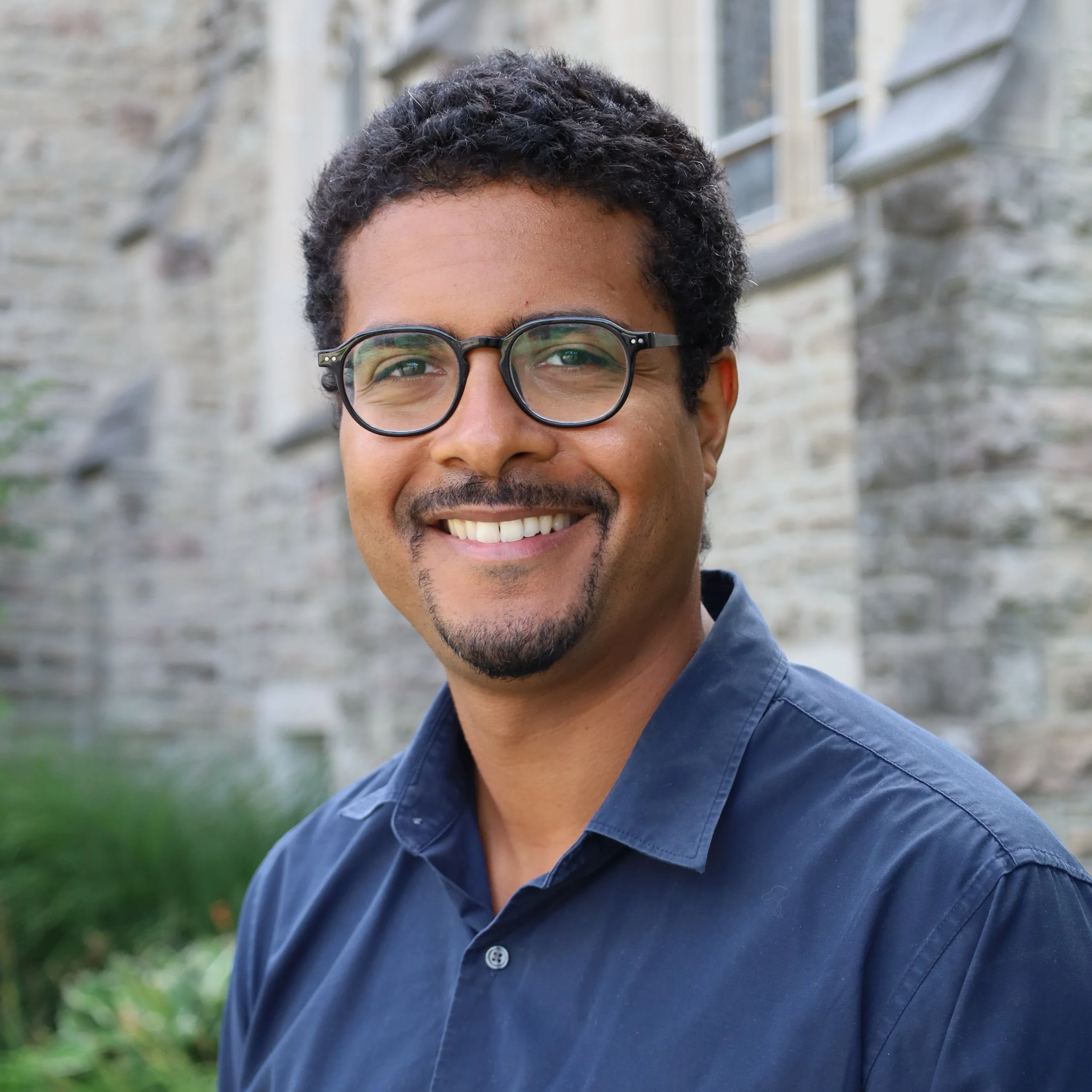 A smiling man with curly black hair, glasses, and facial hair outside in front of an old building with stone walls and large windows.