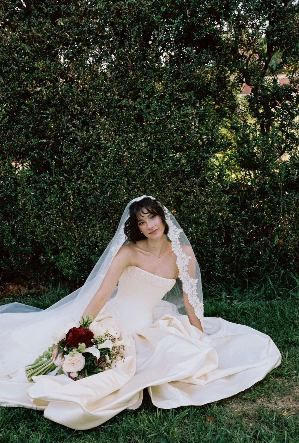 A woman in a wedding dress sitting outdoors on grass with a bouquet of flowers, surrounded by green bushes.