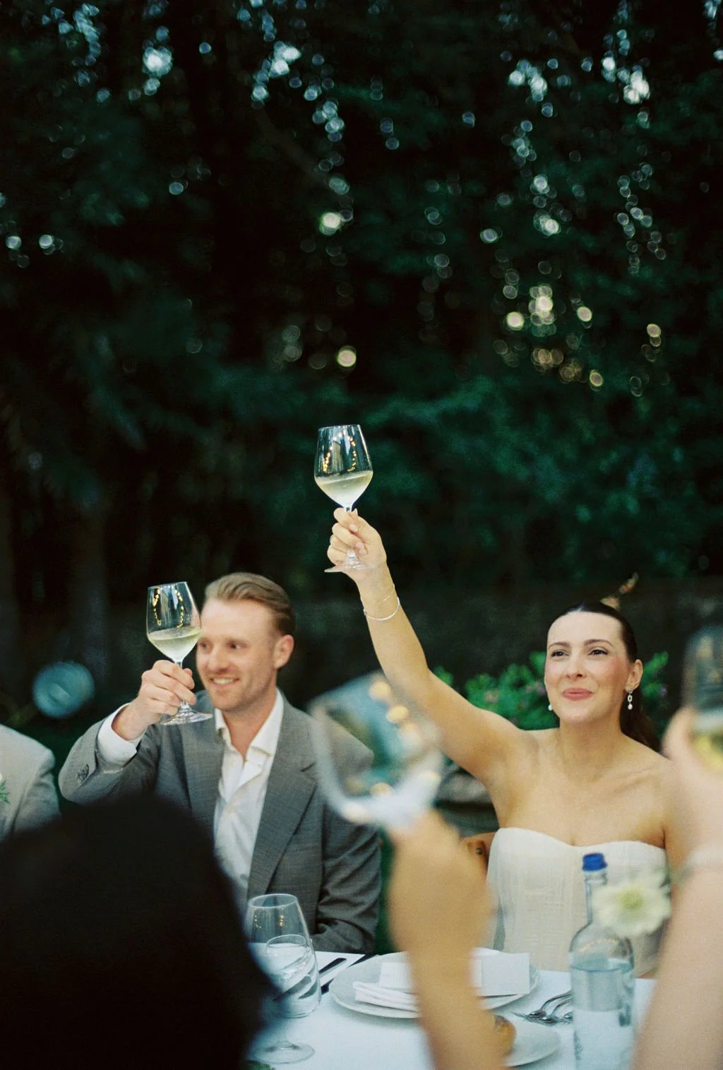 A woman and a man are celebrating, raising their wine glasses in a toast at a dinner gathering outdoors with a lush green background.