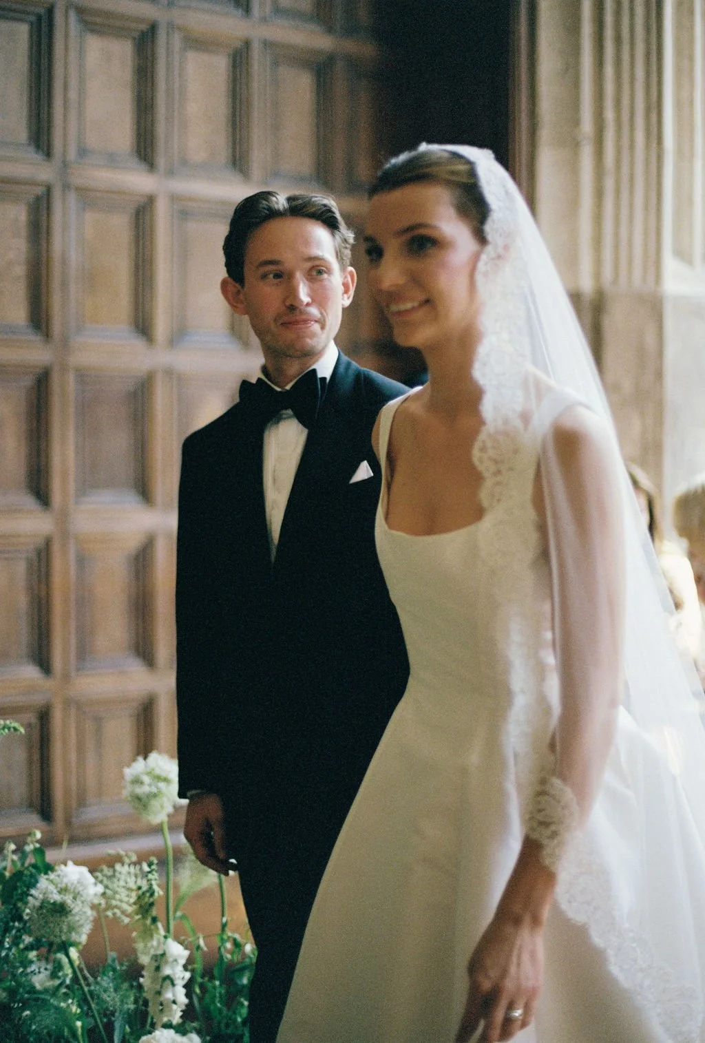 A bride and groom standing together indoors during a wedding ceremony. The bride is wearing a white wedding dress with lace details and a veil, smiling, while the groom is in a black tuxedo with a bow tie, looking at her.