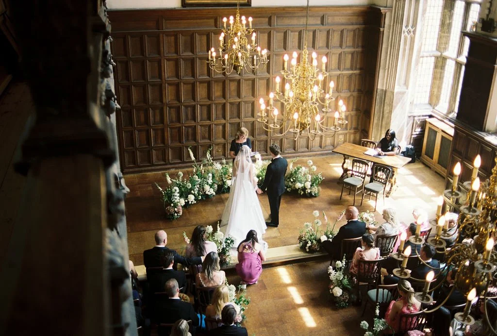 A wedding ceremony taking place in a grand, wood-paneled room with chandeliers and floral arrangements, with the bride and groom standing at the altar facing officiant, surrounded by seated guests.