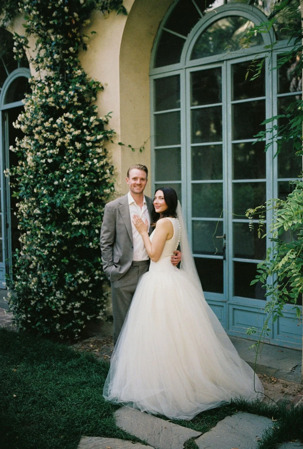 A bride and groom standing outdoors in front of large blue window panes, surrounded by lush green plants and flowers. The bride is wearing a white wedding gown with a veil, and the groom is in a gray suit with an open-collared white shirt.