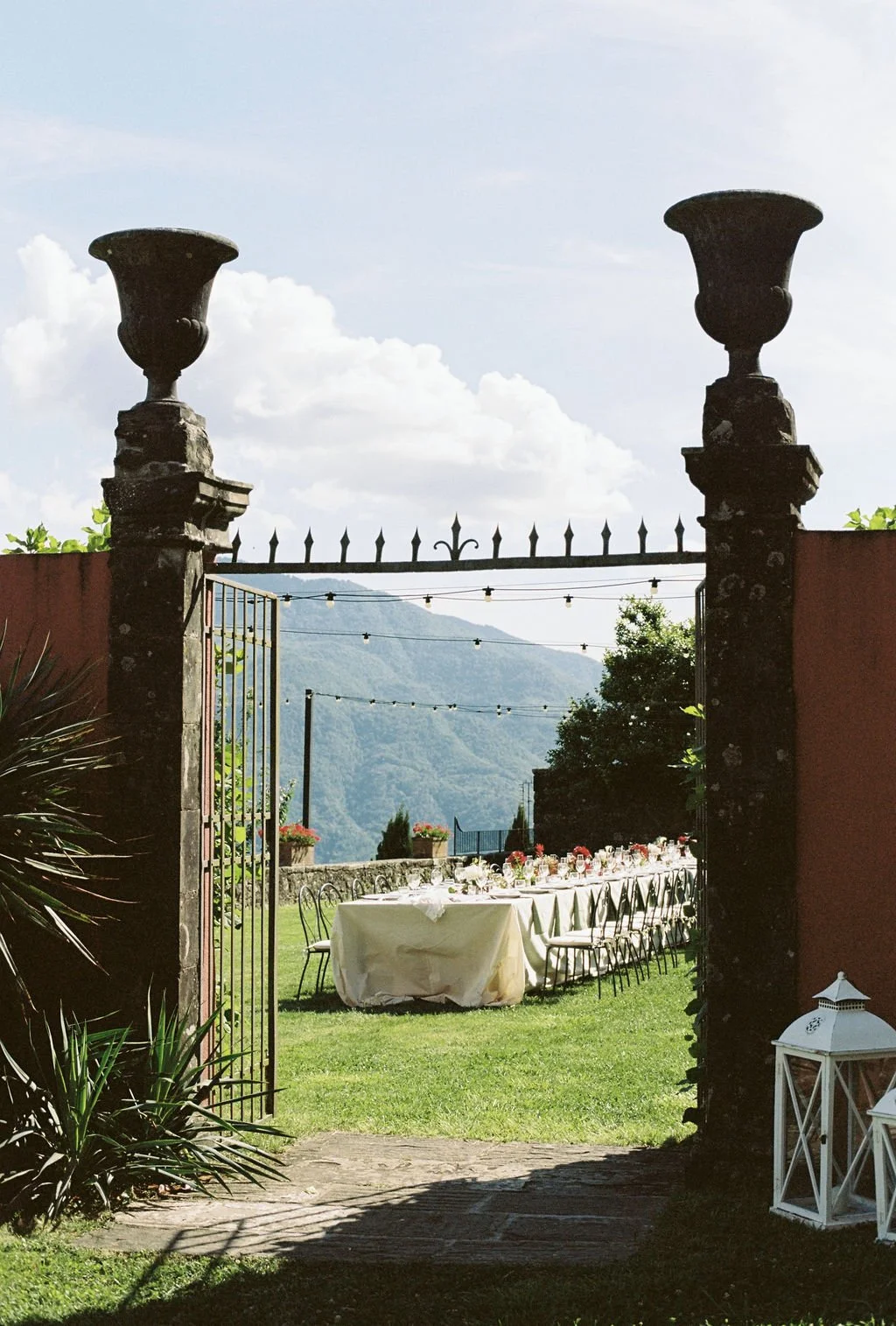 Outdoor dining setup with a long table covered with a cream tablecloth, adorned with candles and flowers, set on a lush green lawn, viewed through an ornate wrought-iron gate with stone columns, with mountains in the background and string lights hang