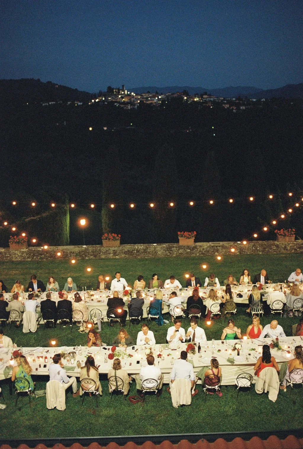 Outdoor dinner party at night with long tables, string lights, and guests seated on chairs, with hills and a town illuminated in the background.