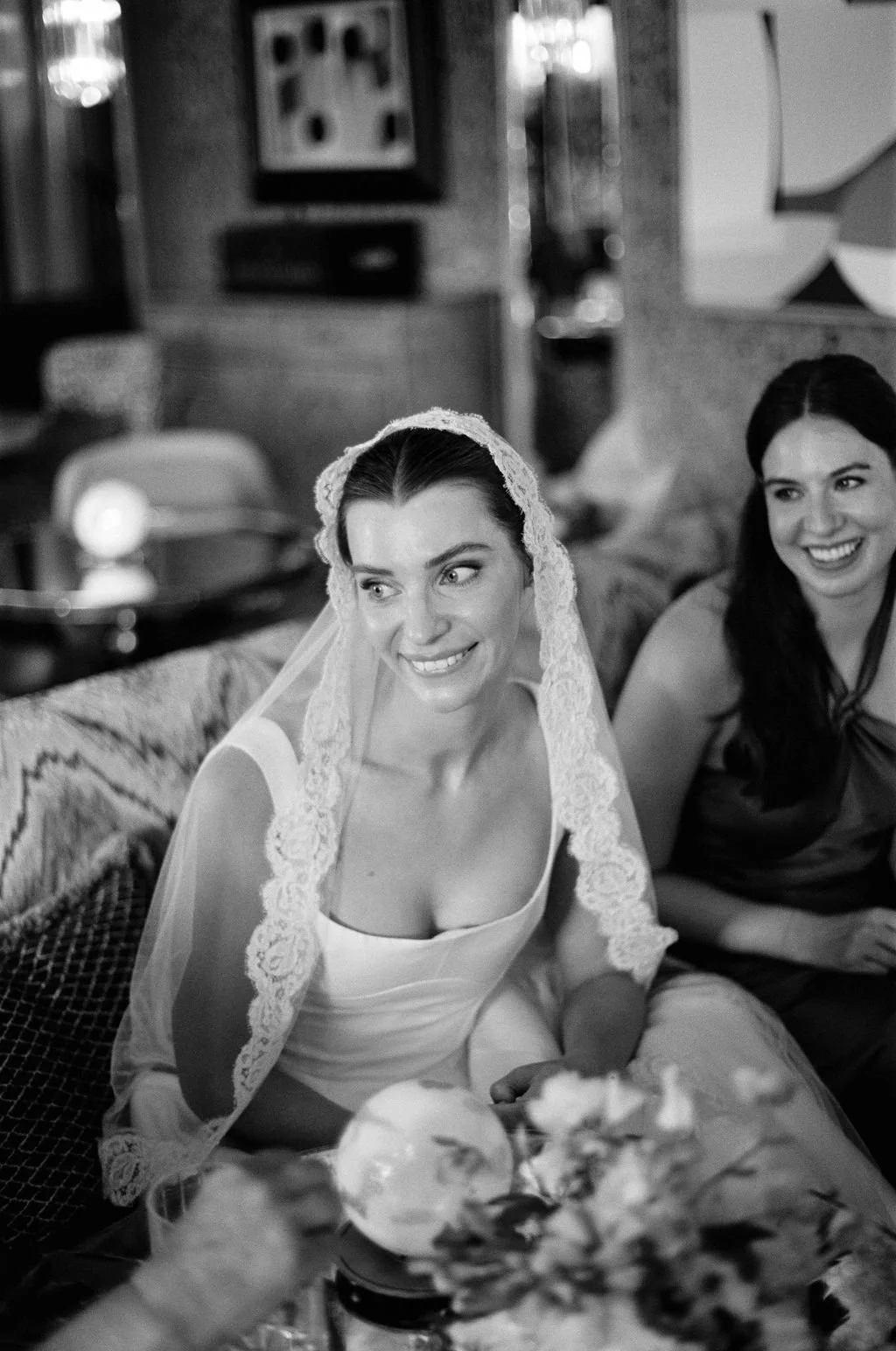 A smiling bride wearing a veil and wedding dress, sitting on a sofa, with a woman next to her, in a cozy indoor setting.