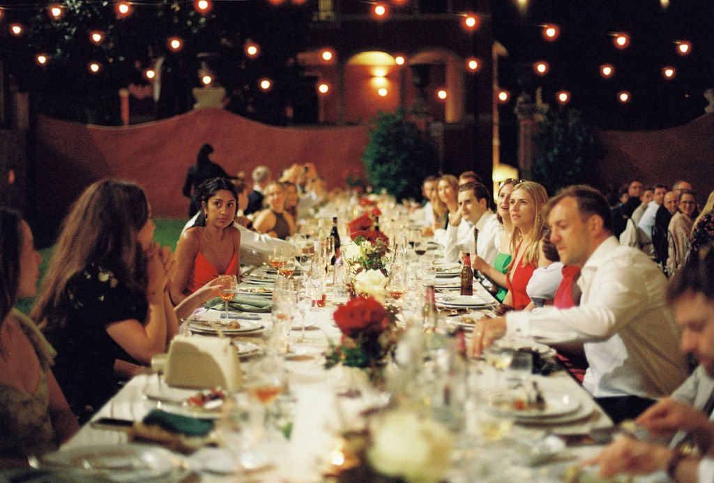 People sitting at a long, decorated dinner table outdoors during evening with string lights overhead.
