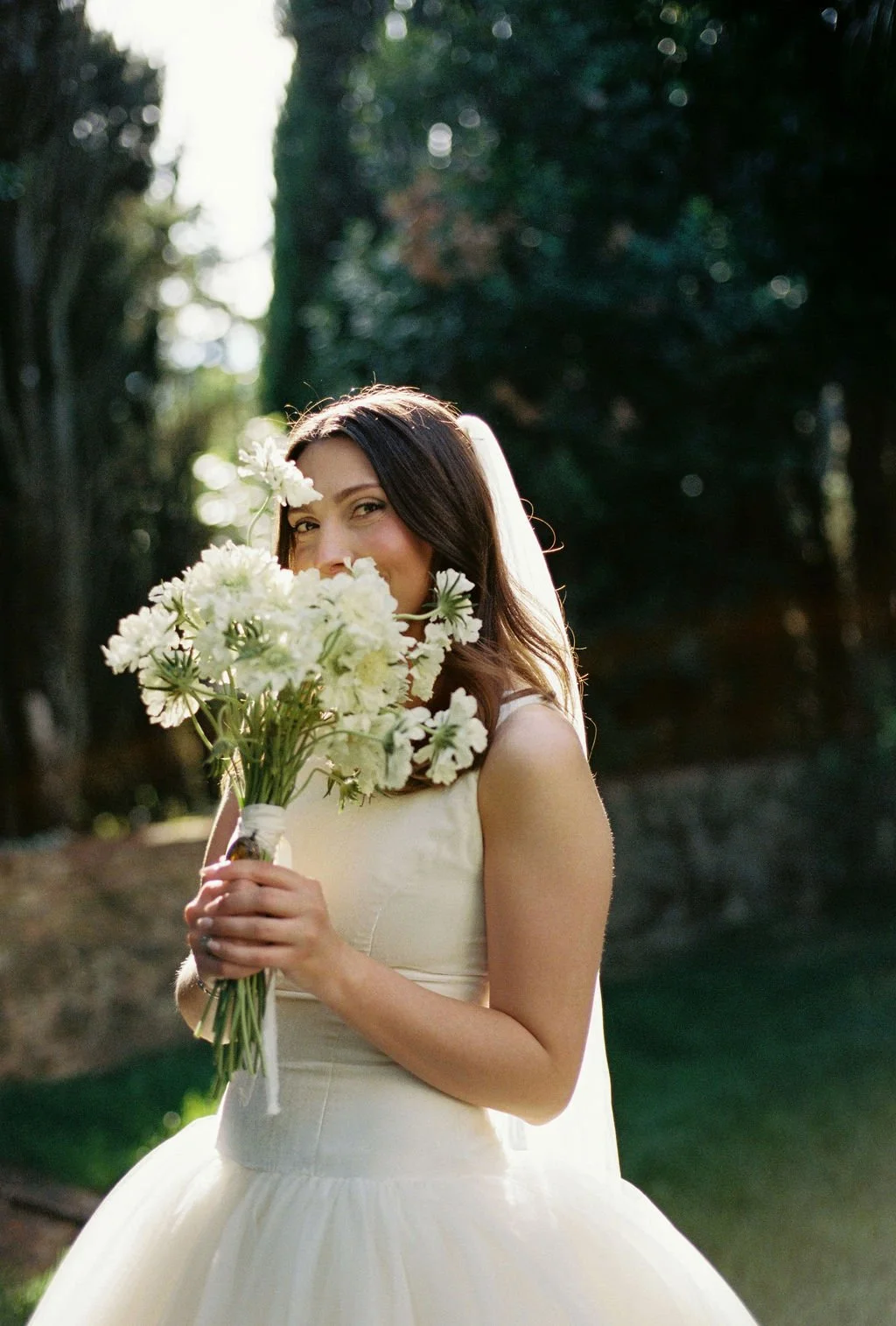 A woman in a white wedding dress holding a bouquet of white flowers, smiling and peeking from behind the bouquet outdoors with trees in the background.