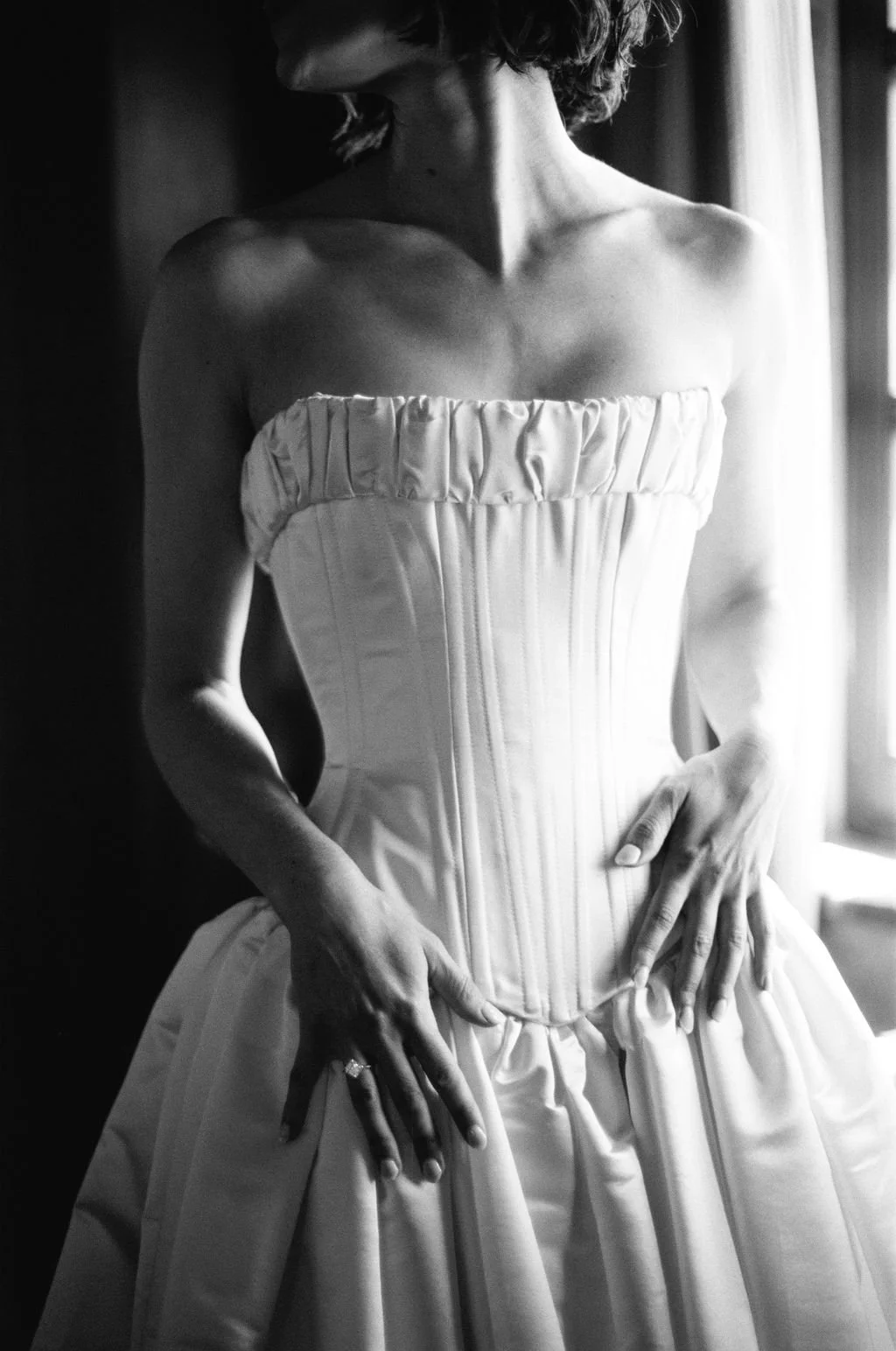 A woman wearing a strapless, corset-style wedding dress, standing beside a window with curtains, with her hands resting on her hips.