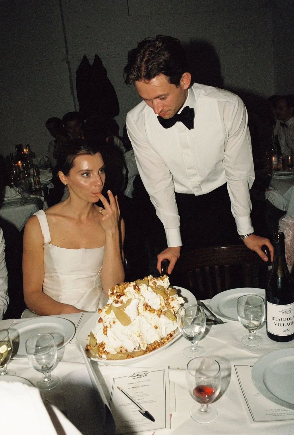 A woman with short dark hair, wearing a white dress, sitting at a table with a large dessert of whipped cream, caramel, and pastry. A man in a white shirt with a black bowtie is leaning over her. The table has glasses, a menu, and a bottle of wine. O