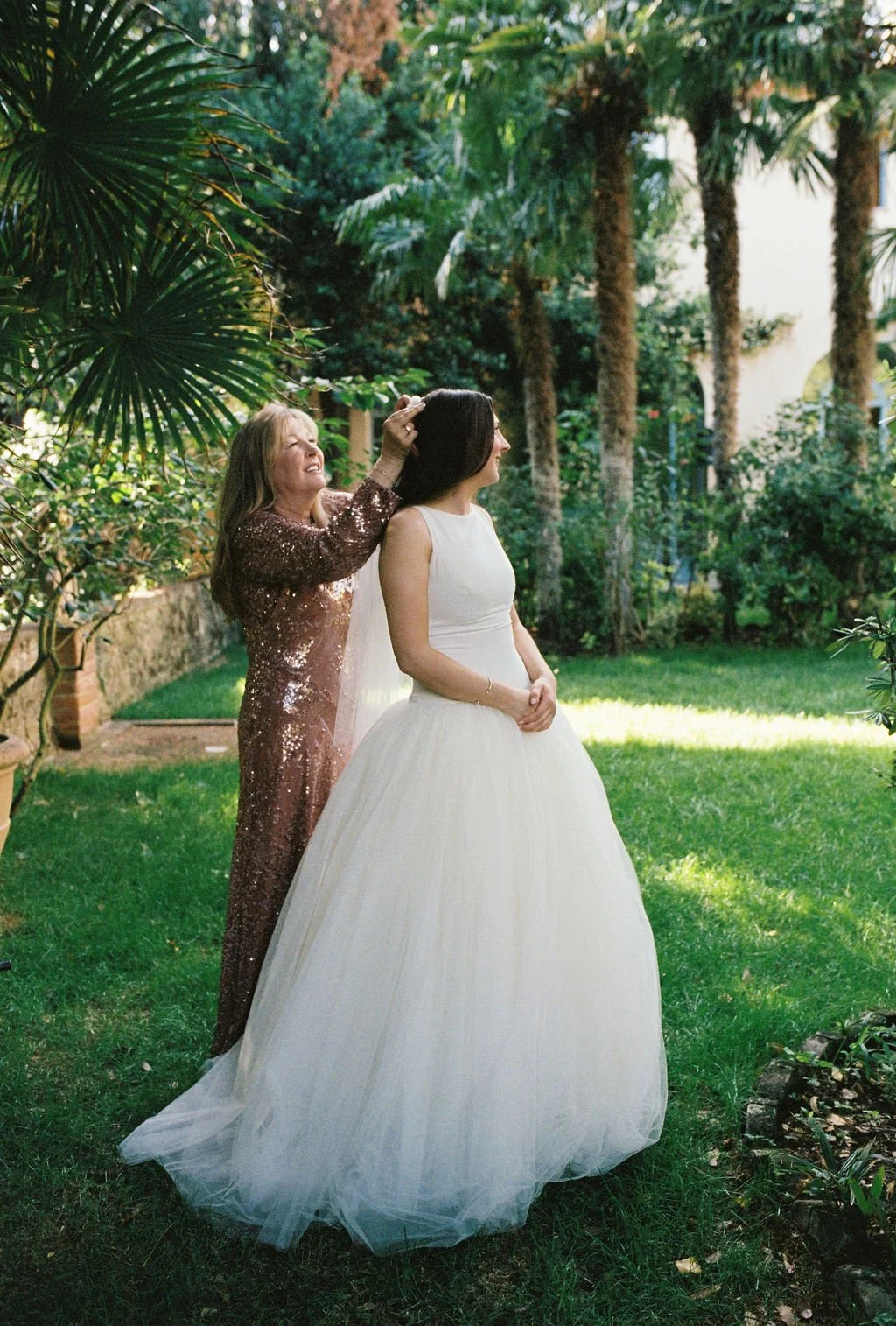 A bride in a white wedding gown standing outdoors in a lush garden, while a woman in a brown, sequined dress adjusts her hair.
