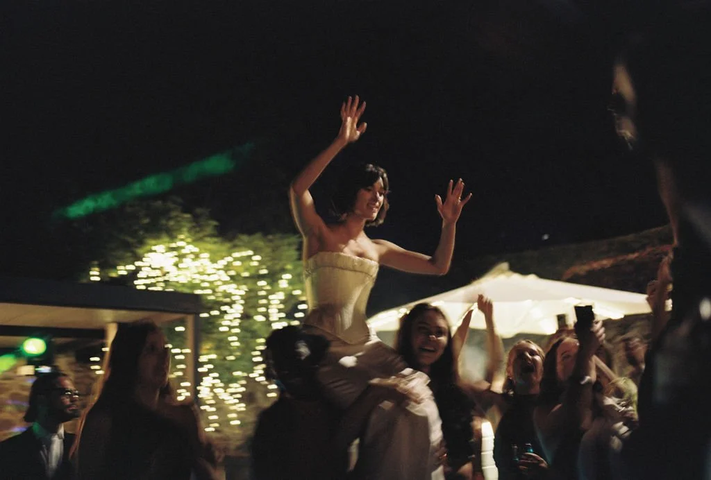 A woman being lifted onto someone's shoulders at a celebration or party outdoors at night, with string lights and a tent in the background.