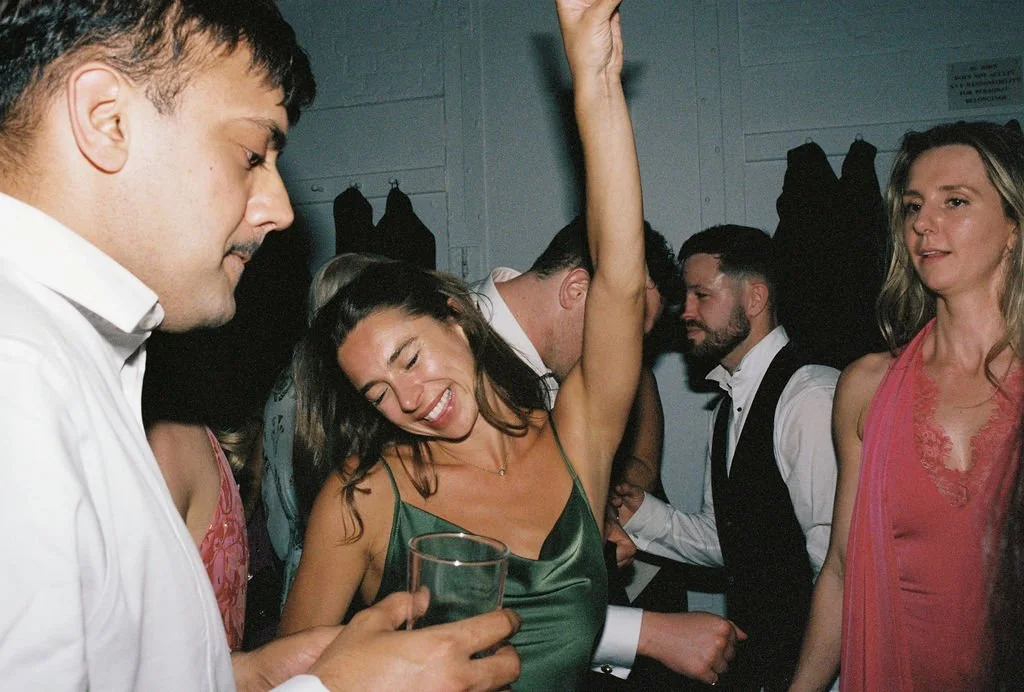 People dancing at a party, a woman in a green dress dancing with her eyes closed and a drink in hand, others surrounding her with relaxed expressions.