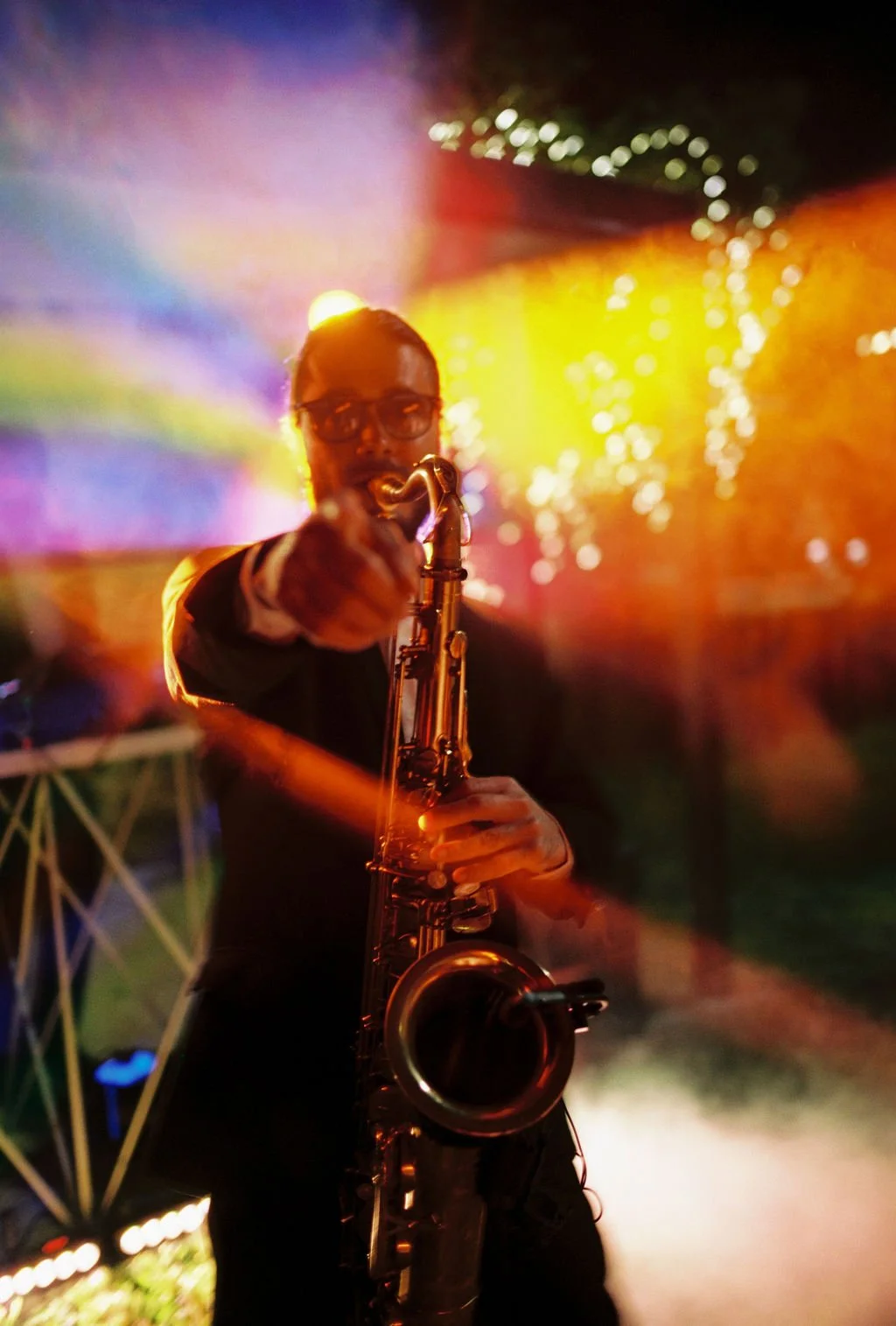 A man wearing glasses and a suit jacket playing a saxophone at a vibrant, colorful party or concert with multicolored lights and smoke in the background.
