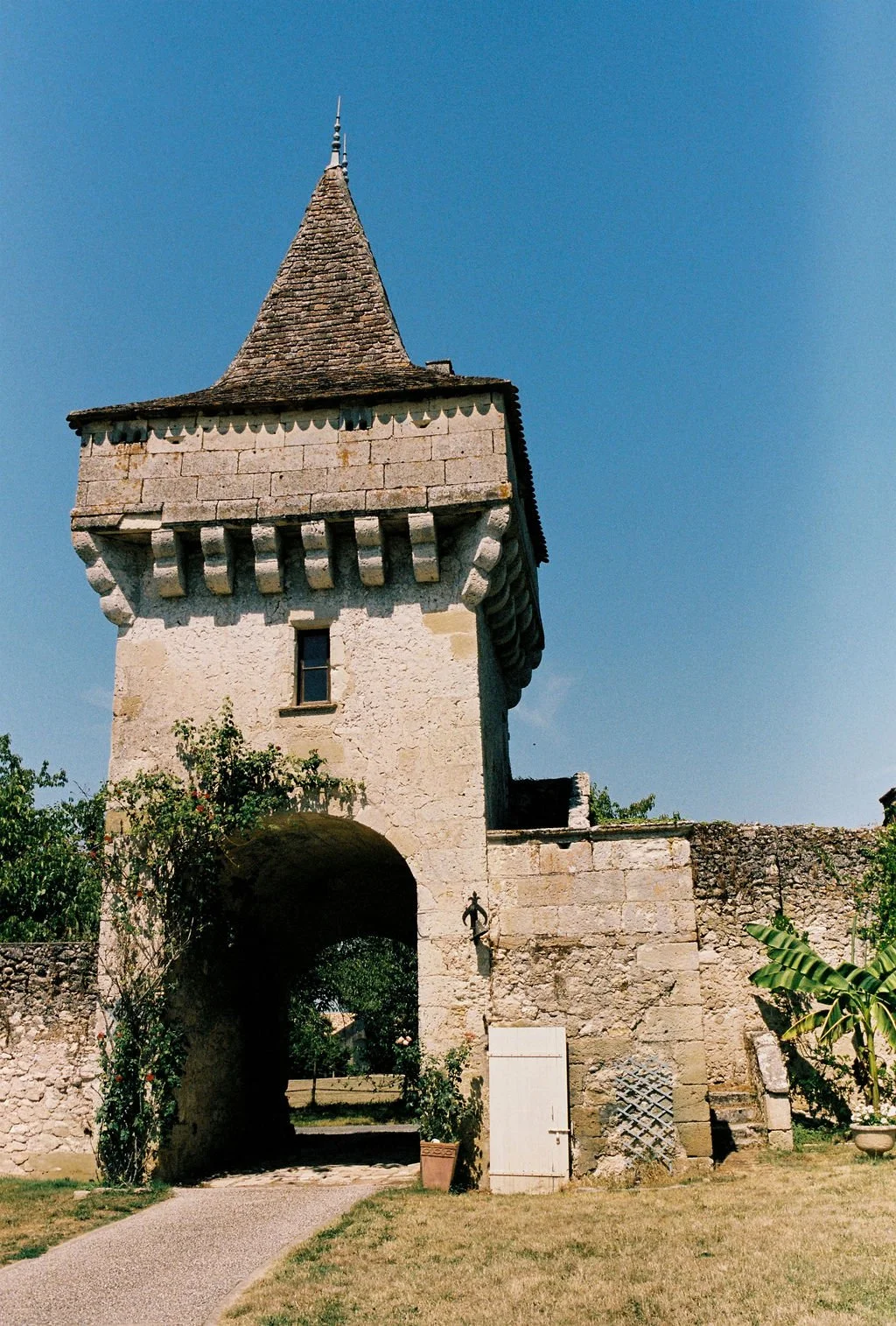 A historic stone tower with a pointed, shingled roof, arched entrance, small window, and decorative stone details, set against a clear blue sky and surrounded by greenery.