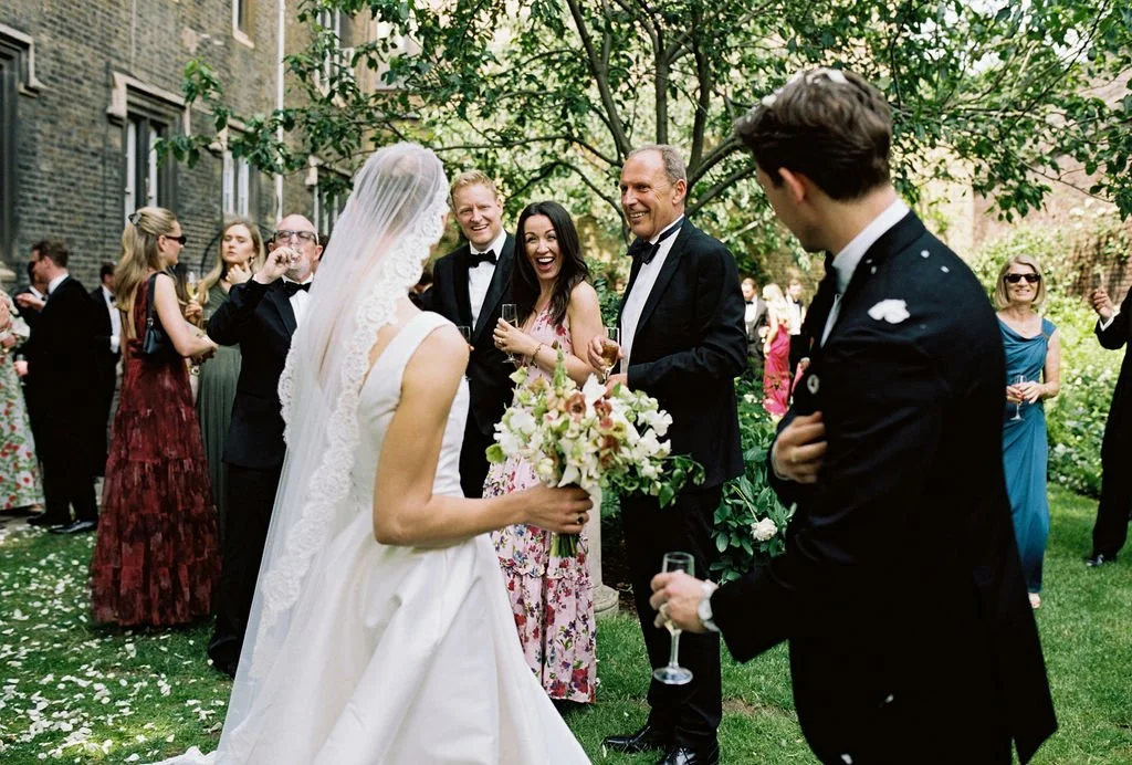 Bride holding a bouquet, smiling and talking with guests at an outdoor wedding reception, surrounded by guests in formal attire, under a tree with green foliage.