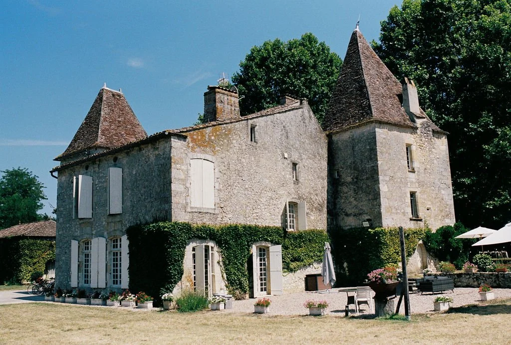 A historic stone castle with two conical towers and white shutters, surrounded by greenery and outdoor seating with umbrellas
