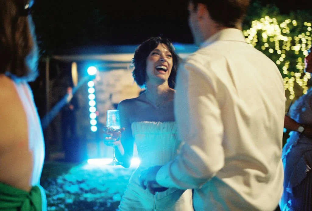 A woman with short dark hair smiling and laughing while holding a glass, talking to a man at an outdoor evening party with string lights in the background.