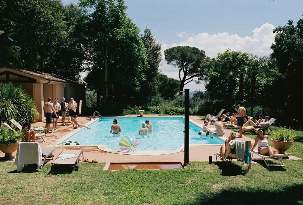 People swimming and relaxing by a backyard swimming pool on a sunny day, surrounded by lush green trees and garden furniture.