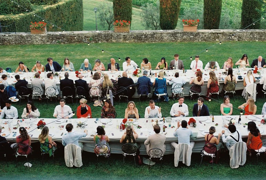 Long outdoor banquet table set up for a formal event, with guests seated, decorated with flowers and string lights, surrounded by trees and greenery.