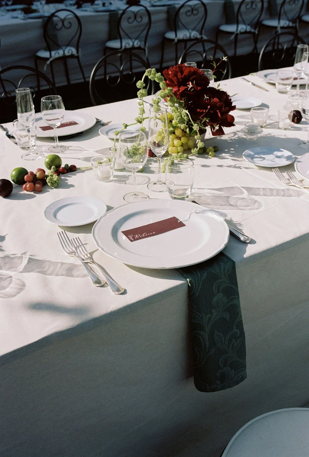 Elegant dining table set outdoors with white tablecloth, multiple wine glasses, plates, silverware, a floral centerpiece with red and green flowers, and a place card labeled 'Rebecca' at the center.