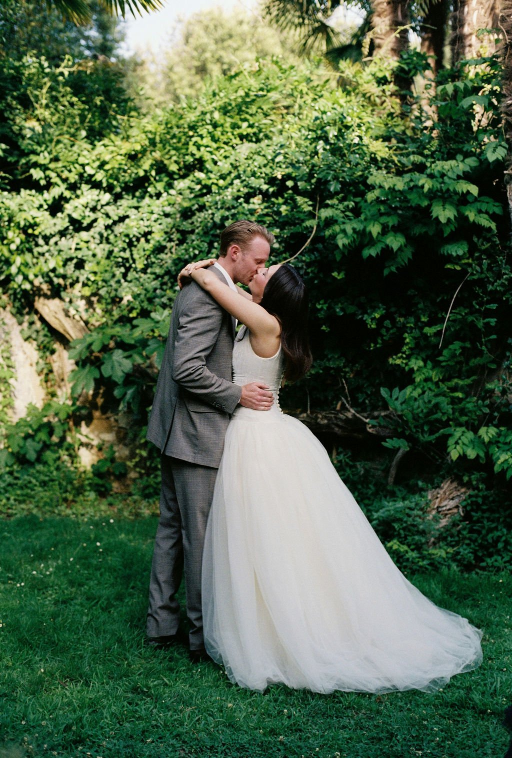 A couple in wedding attire sharing a kiss outdoors with lush green foliage in the background.