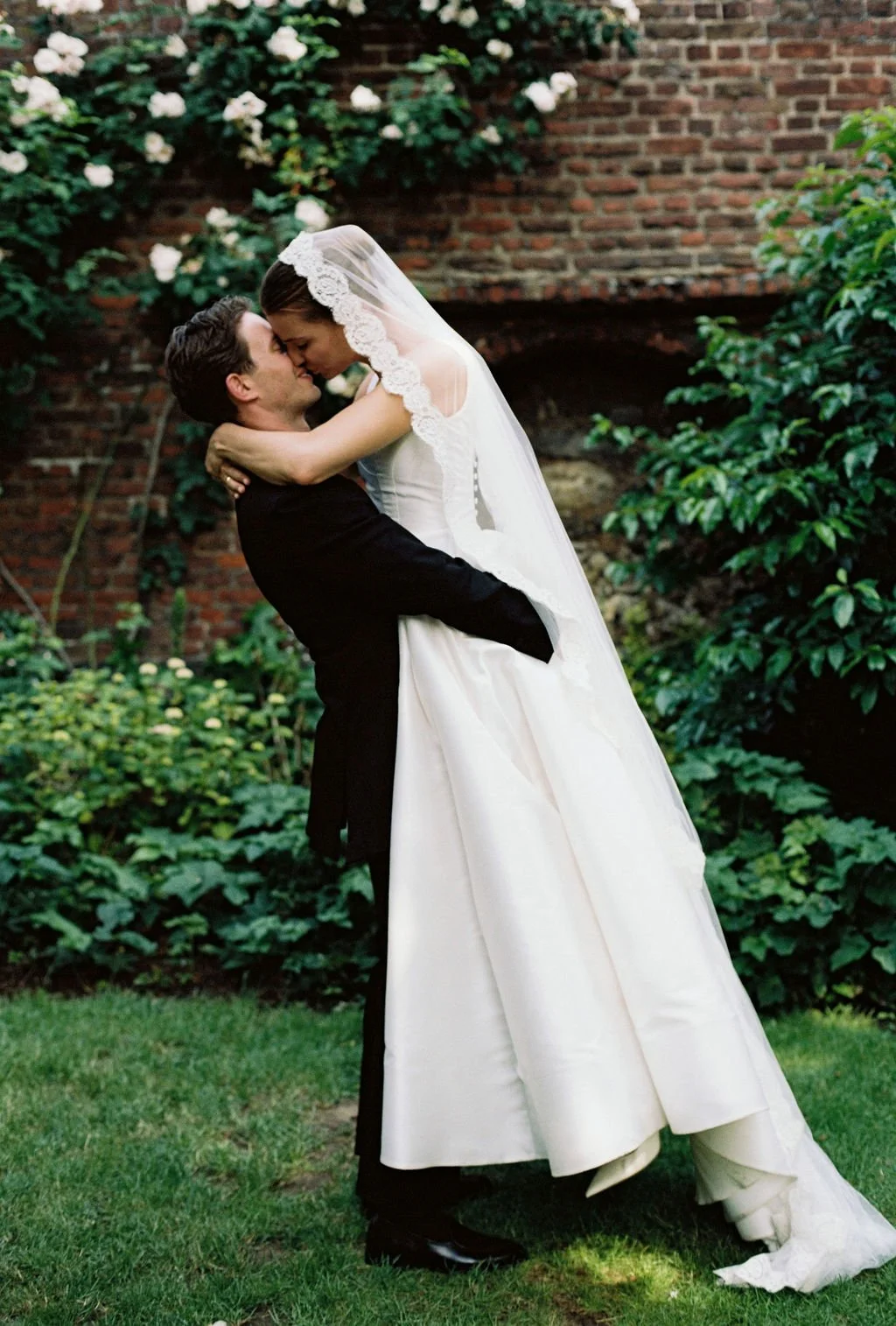 A bride and groom in wedding attire embrace outdoors in front of a brick wall and green foliage.