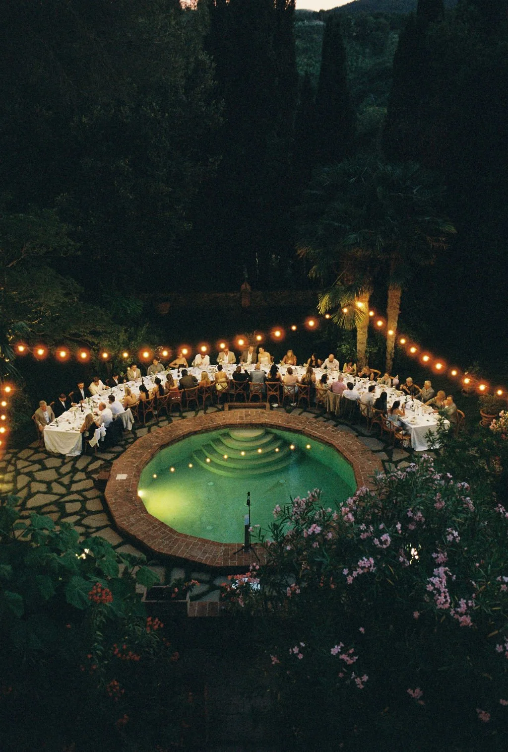 An outdoor dinner party at night around a brick-lined pool with steps, with long tables and chairs, string lights overhead, surrounded by trees and foliage.