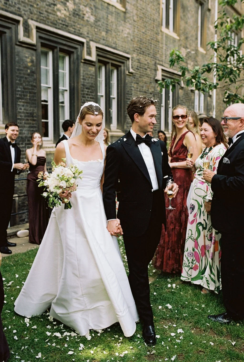 Bride and groom holding hands, wedding celebration outdoors, surrounded by guests in formal attire, bride holding bouquet, groom in tuxedo, guests smiling and holding champagne glasses.