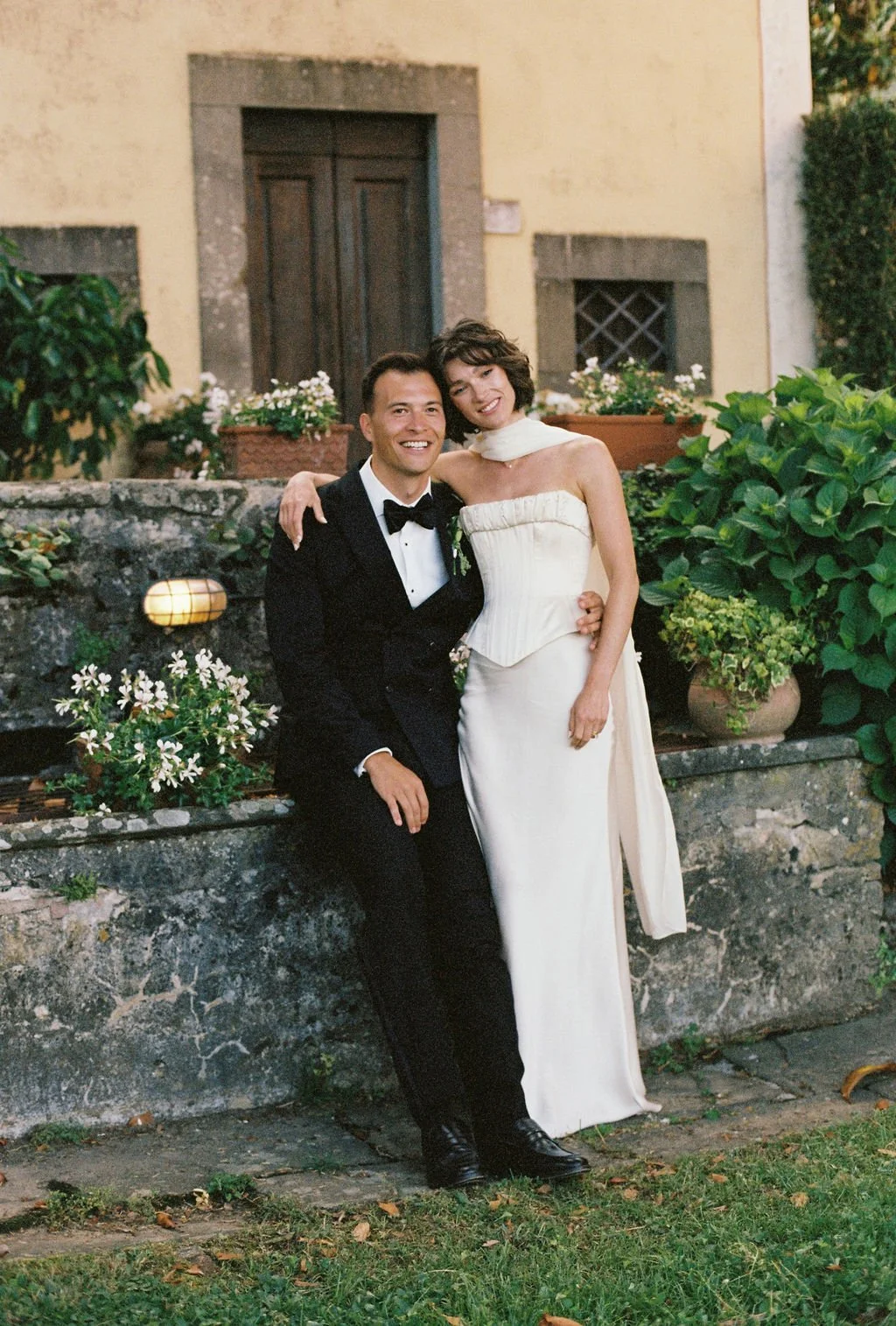 A smiling couple, man in a tuxedo and woman in a white wedding dress, pose together outdoors in front of a stone ledge with flowers and greenery, at a wedding or special event.