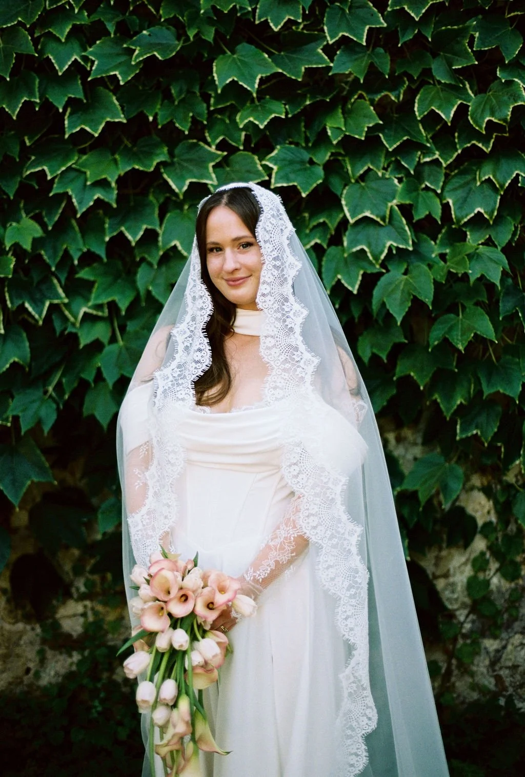 A young woman in a white wedding dress and lace veil holding a bouquet of light pink flowers, standing in front of green ivy leaves.