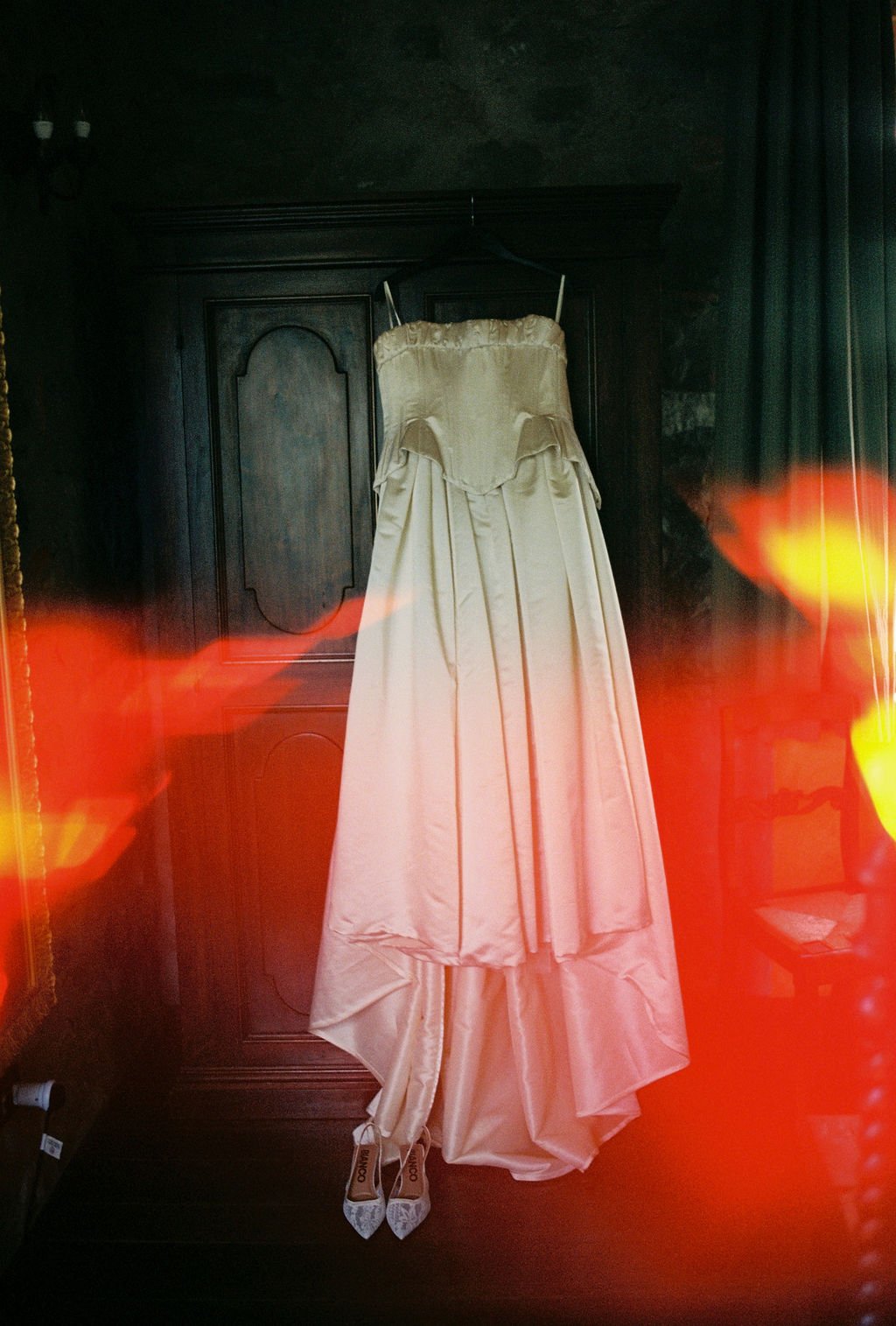 A wedding dress hangs on a black wooden wardrobe. The train of the dress is pooled on the floor with a pair of white high-heeled shoes underneath. The setting appears to be a dimly lit room with dark walls and curtains, with red and yellow light refl