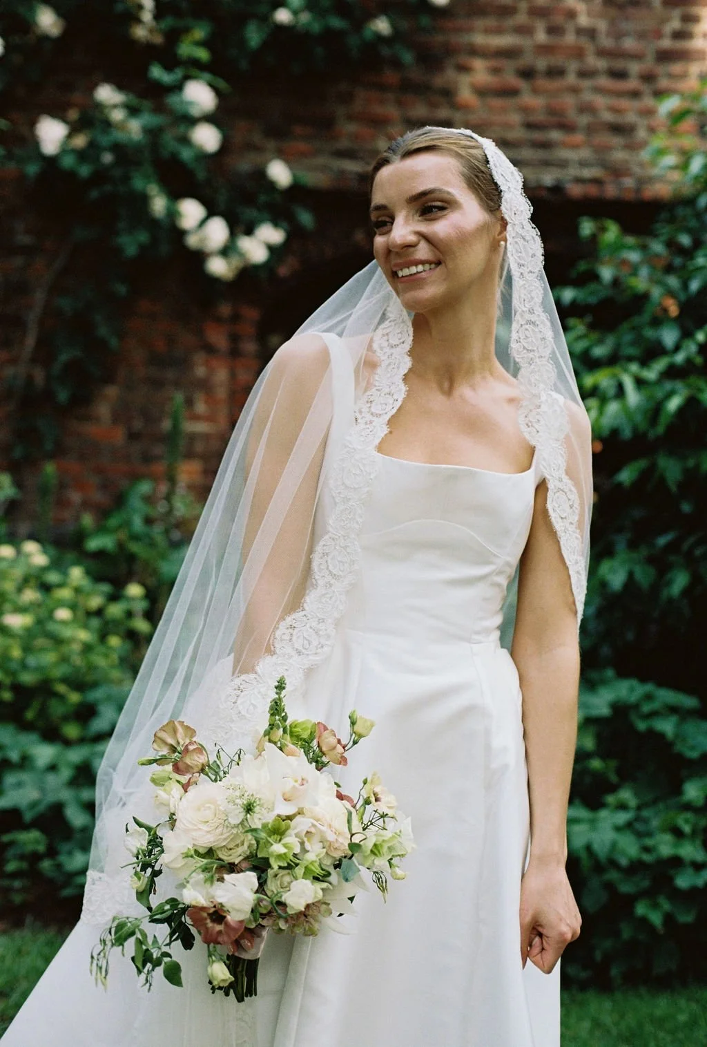 A bride wearing a white wedding gown, lace veil, and holding a bouquet of white and pink flowers, standing outdoors in front of green foliage and a brick wall.