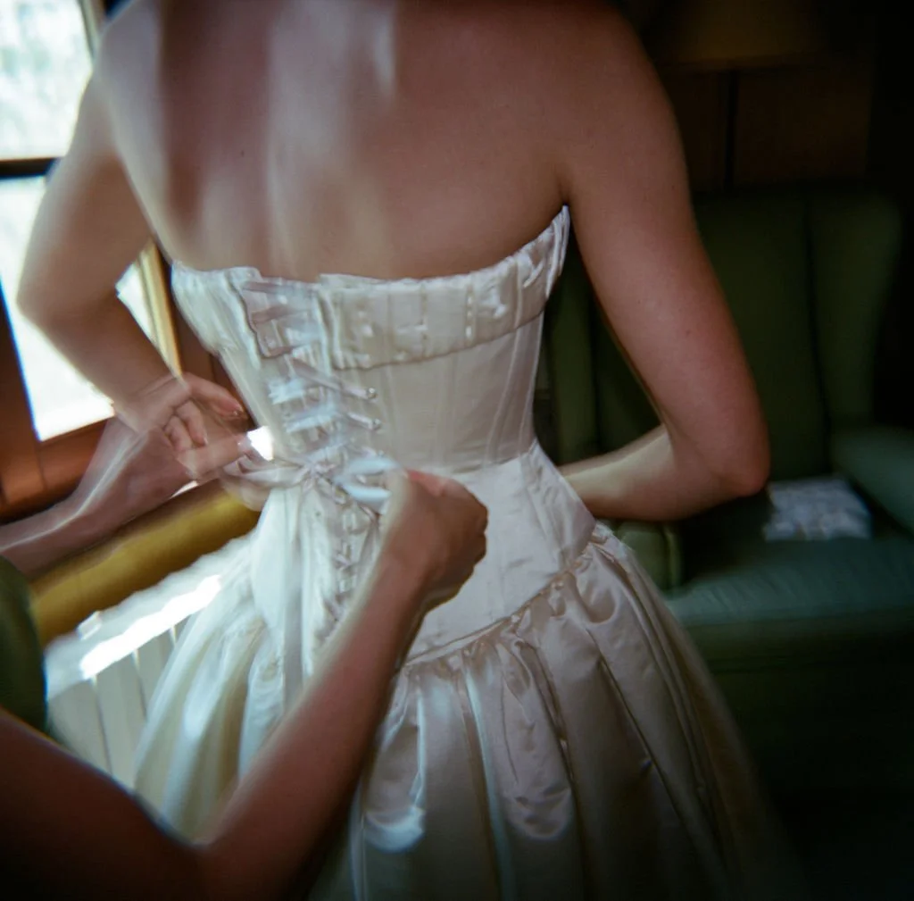 A woman wearing a white satin wedding dress being helped into her gown by another person.