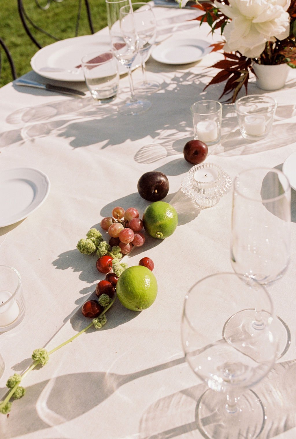 A table set outdoors with white tablecloth, glassware, plates, and napkins. Centerpiece includes flowers and a fruit arrangement of grapes, limes, cherries, and plums, with candles and candle holders.