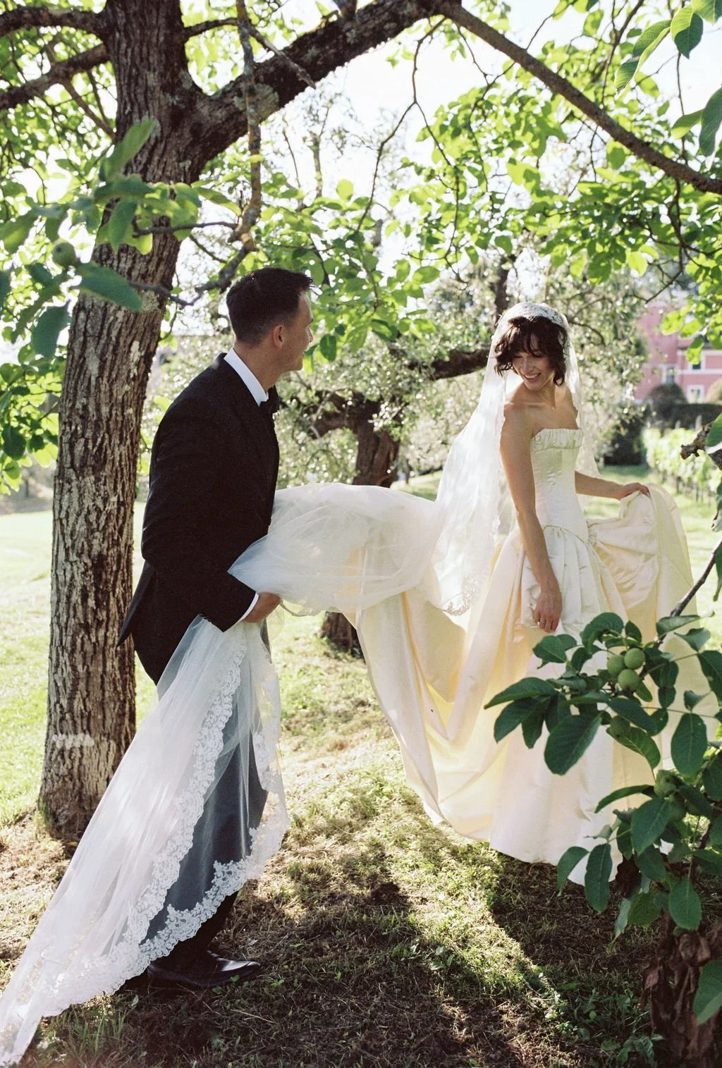 A bride and groom in wedding attire standing outdoors under a tree, with the groom holding the bride's dress as they smile at each other.