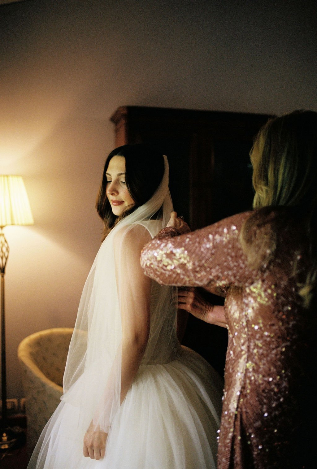 A bride in a white wedding dress with a veil is being assisted by a woman in a sequined pink dress as she gets ready for her wedding in a softly lit room.