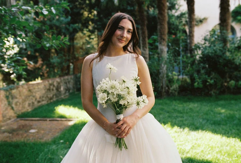 Young woman in a wedding dress holding a bouquet of white flowers in a garden setting.