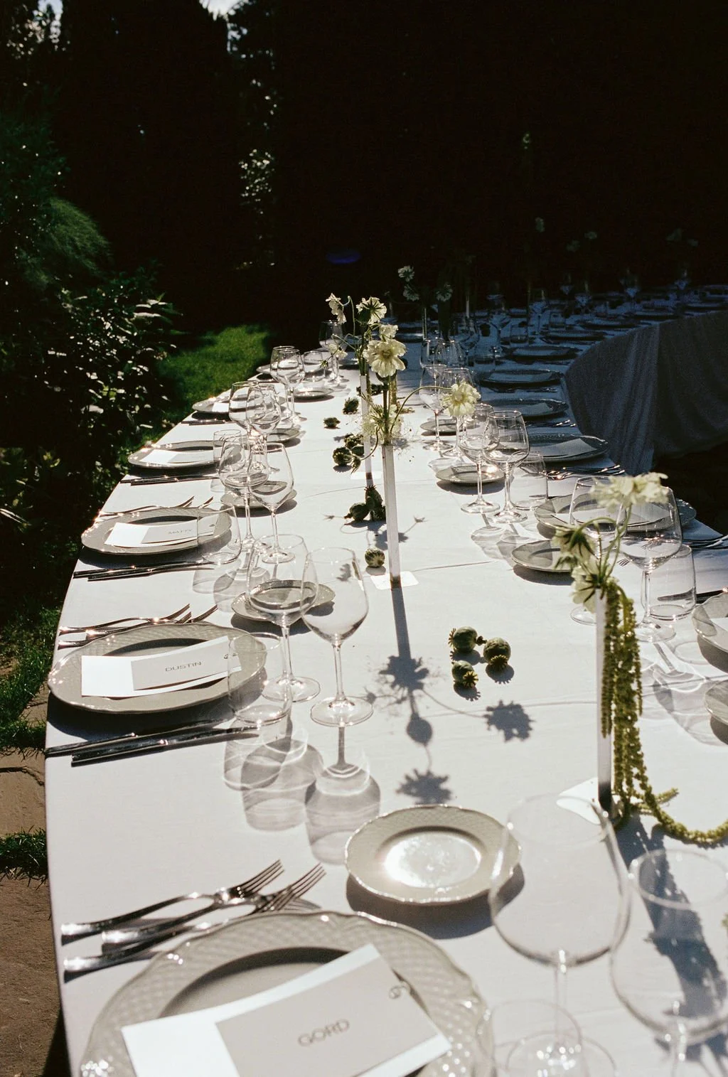 Long outdoor dining table set for a formal event with white tablecloth, flower centerpieces, and arranged glassware and plates, illuminated by sunlight with greenery in the background.