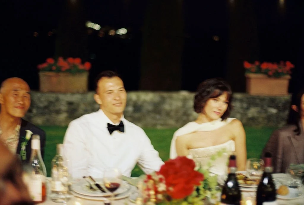 Group of people sitting at a dinner table outdoors at night, with drinks and flowers on the table.