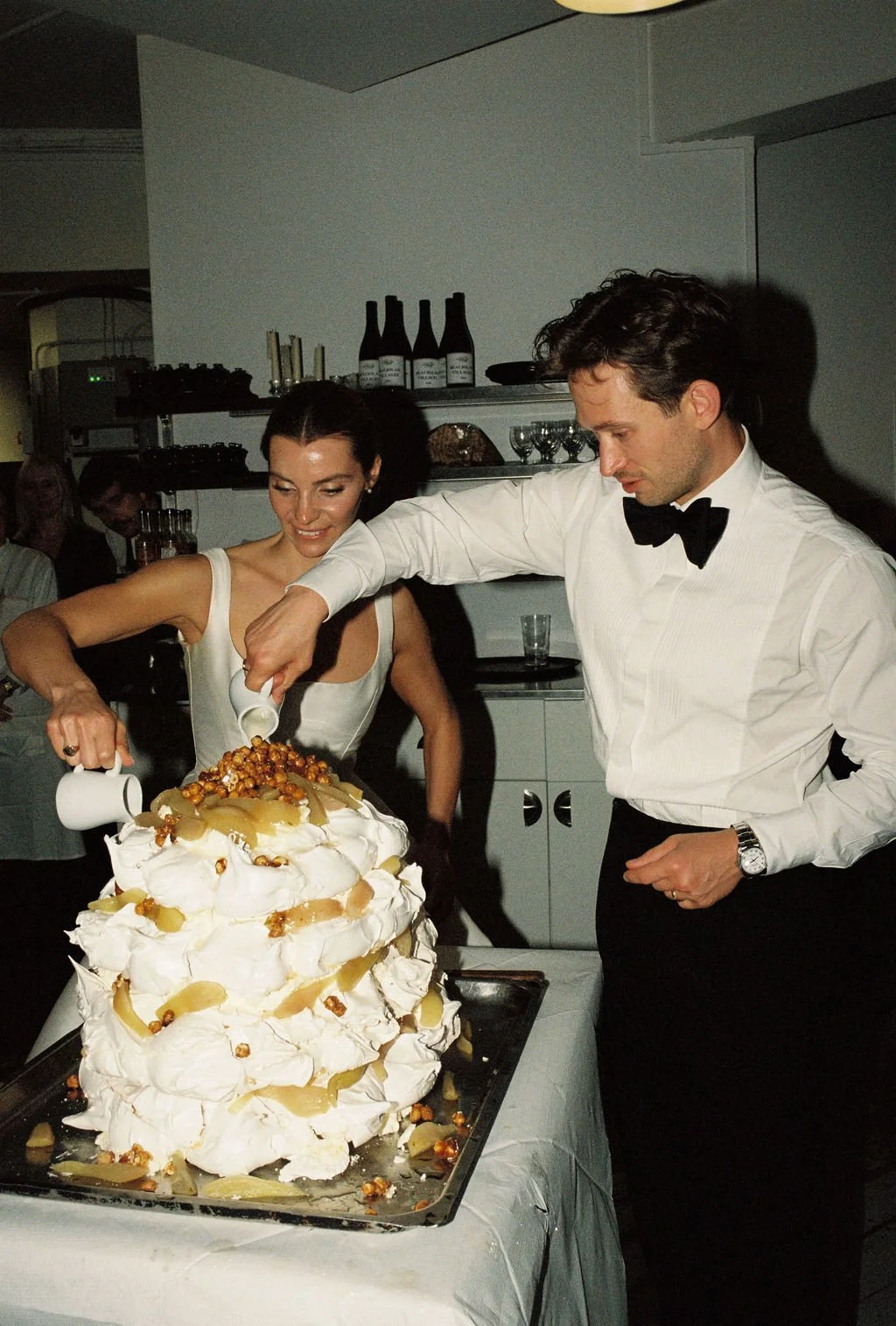 A wedding cake with multiple layers decorated with meringue, caramel popcorn, and pear slices is being cut by a bride and groom during their wedding reception.