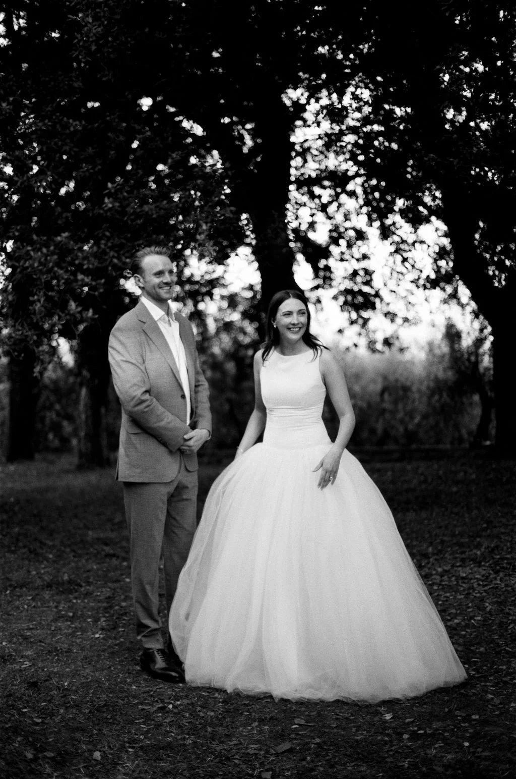 Black and white photo of a bride and groom outdoors, standing on a dirt path under large trees. The groom is dressed in a light-colored suit and the bride in a sleeveless, full-length white wedding gown. Both are smiling and looking away from each ot
