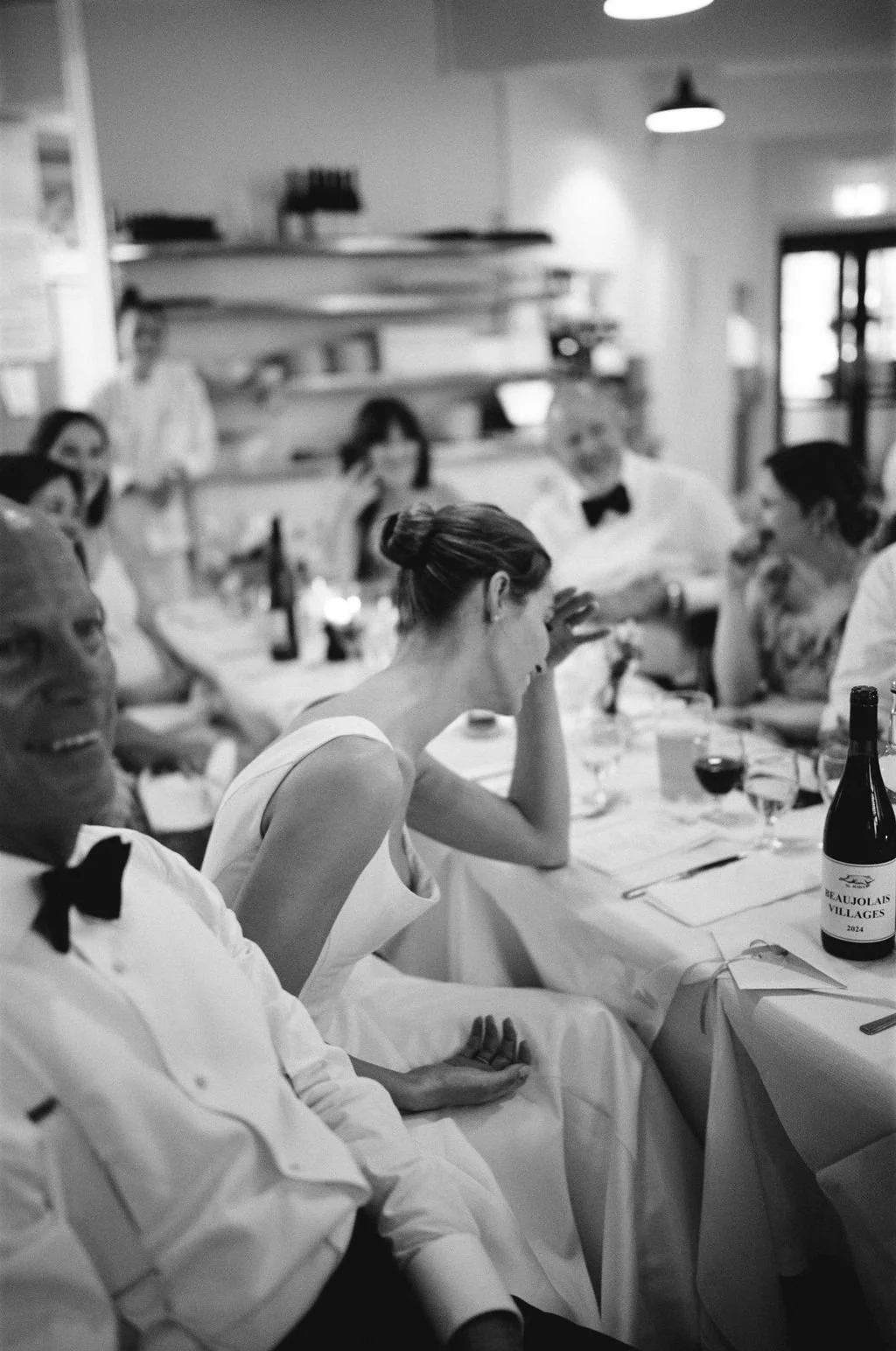 Black and white photo of people dining at a formal event, some wearing white dresses and tuxedos, sitting at a banquet table with wine glasses and bottles, in a restaurant or banquet hall setting.