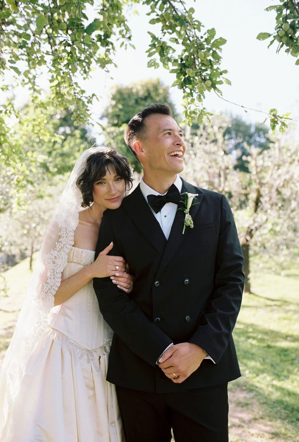 A bride and groom standing outdoors under a tree, smiling and posing for a wedding photo.