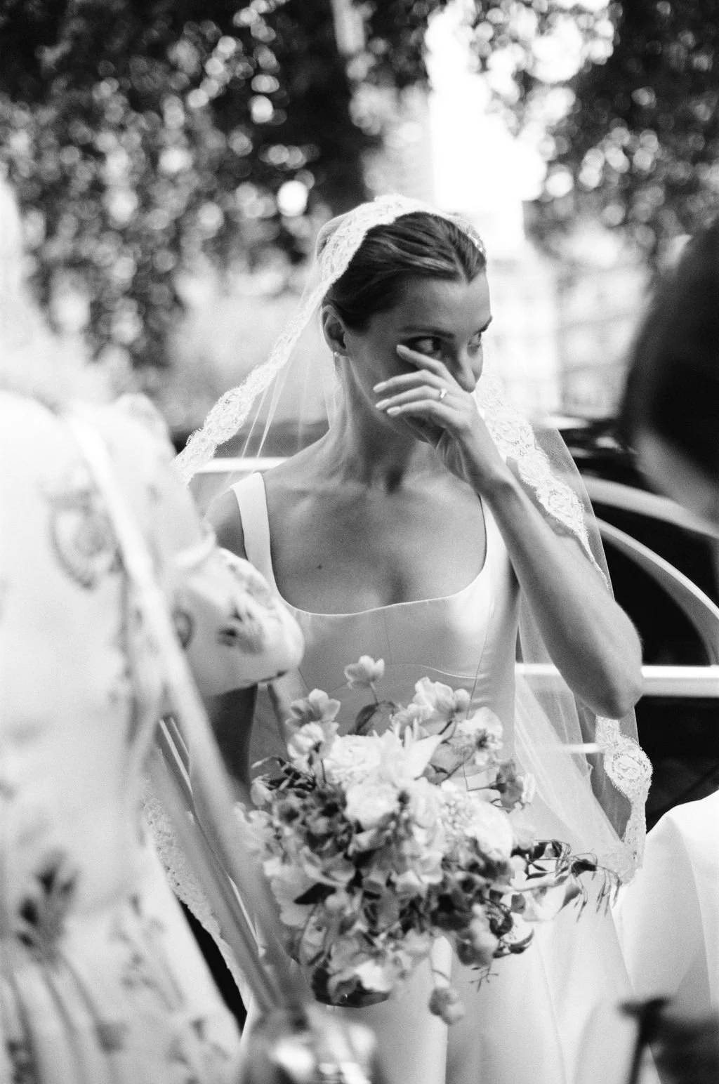 A bride wearing a wedding dress and veil, holding a bouquet, wiping her eyes with her hand, during a wedding ceremony.