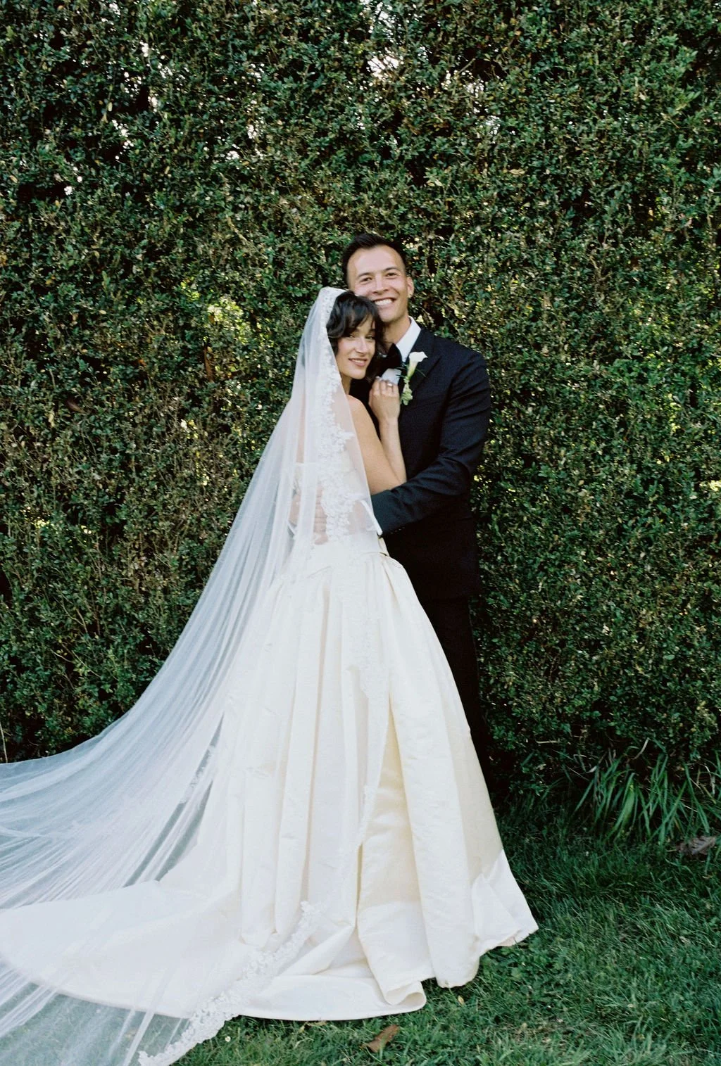 A newlywed couple standing in front of a green hedge, with the bride in a white wedding gown and veil, and the groom in a black tuxedo, smiling and embracing.