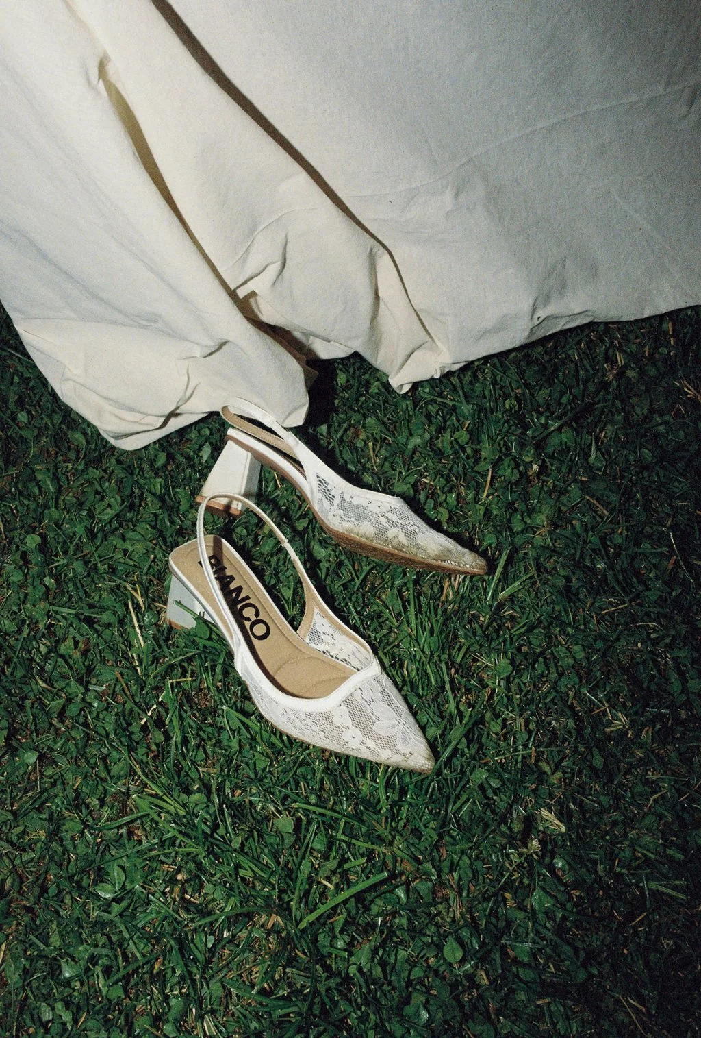 White lace high heels on green grass with a white fabric in the background.
