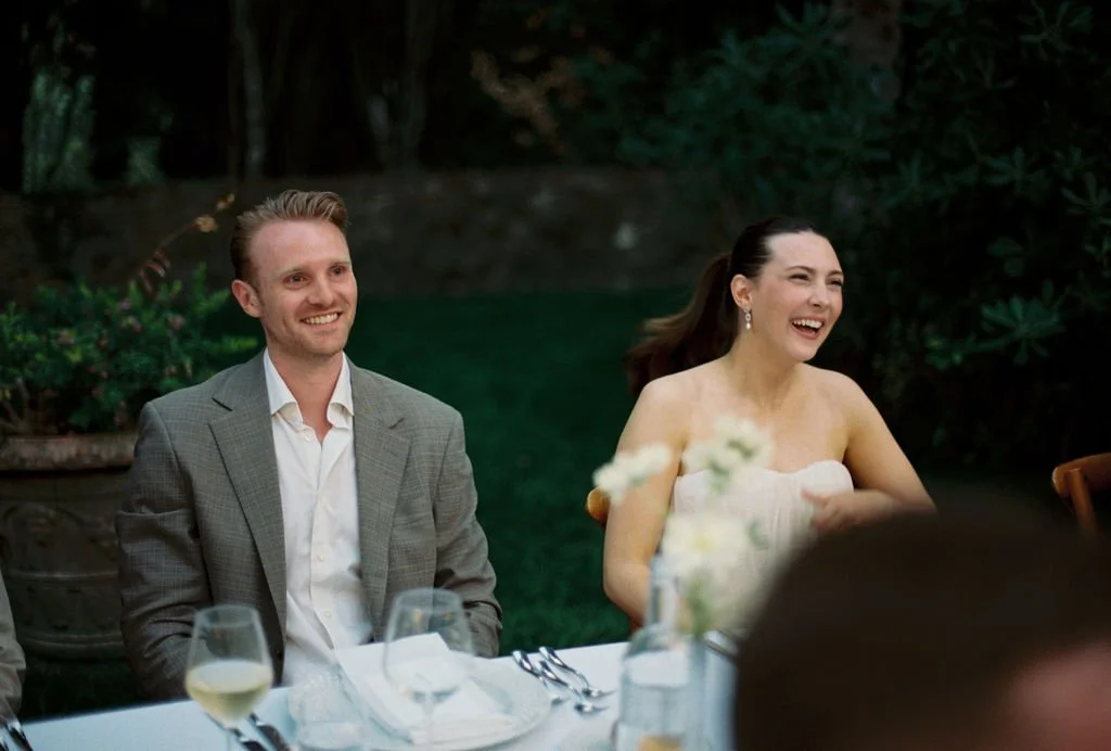 A man in a gray suit and a woman in a white dress are sitting at a table outdoors, smiling and laughing during a wedding reception.