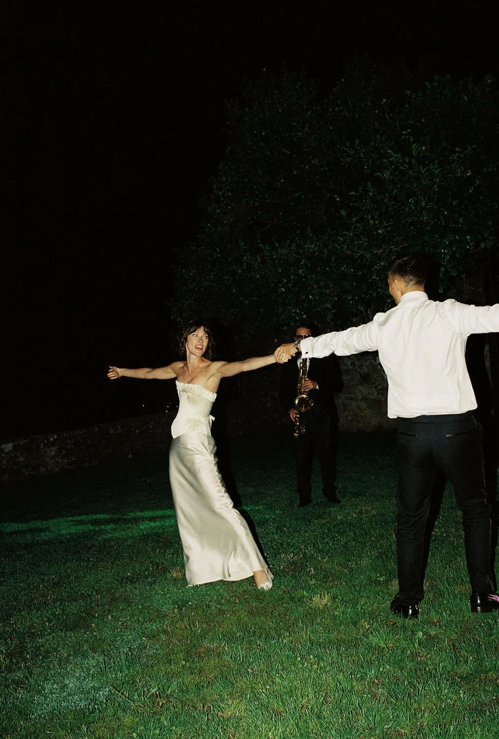 Woman in a white dress dancing outdoors at night while a man in a white shirt and black pants holds her hand. A musician with sunglasses and a saxophone observes in the background.