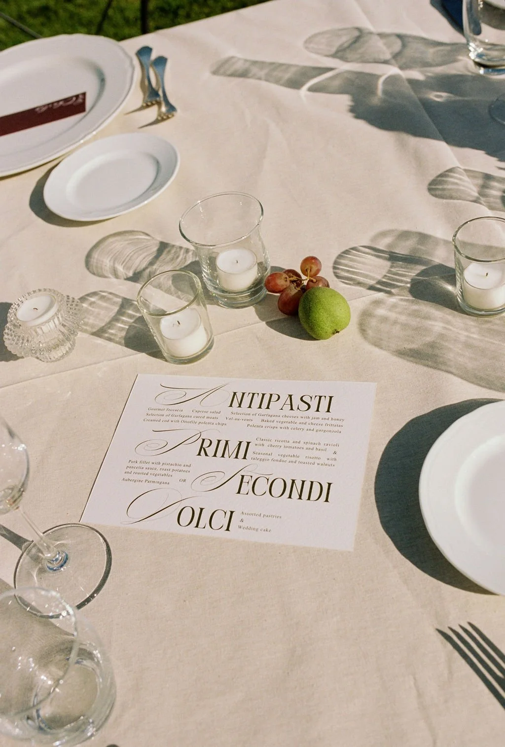 A table setup for a meal with wine glasses, candles, a menu, a small bunch of grapes, a lime, and white plates with silverware, on a cream-colored tablecloth.
